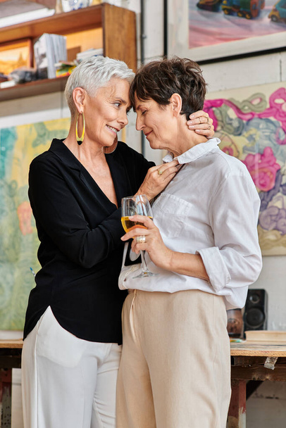 κομψή μεσήλικη γυναίκα αγκαλιάζει λεσβία σύντροφο με ποτήρι κρασιού στο στούντιο σύγχρονης τέχνης - Φωτογραφία, εικόνα