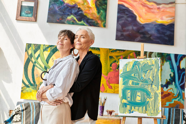 stylish and happy lesbian artists embracing with closed eyes near creative paintings in workshop - Photo, Image