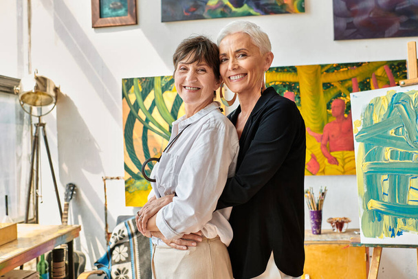 módní a šťastní lesbičtí malíři objímající a dívající se na kameru v blízkosti kreativních obrazů ve studiu - Fotografie, Obrázek