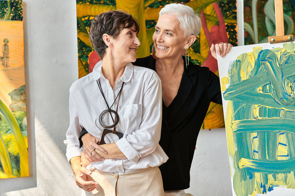 utalentowane dojrzałe lesbijskie artyści obejmujący i uśmiechając się do siebie w pobliżu obrazów w warsztacie - Zdjęcie, obraz