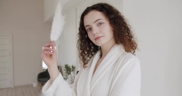 Portrait of young beautiful woman with nude makeup holding playing with feather on face. Beautiful Young Woman with Clean Fresh Skin. Beautiful woman enjoying smooth skin condition at home and smiling - Footage, Video