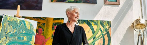 trendy volwassen vrouwelijke kunstenaar glimlachend en wegkijkend in de buurt van kleurrijke schilderijen in kunststudio, banner - Foto, afbeelding