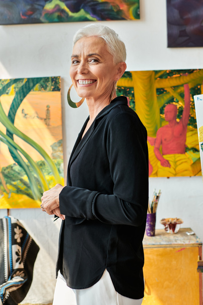 stilvolle und fröhliche Künstlerin mittleren Alters blickt in die Kamera in der Nähe kreativer Gemälde im Atelier - Foto, Bild