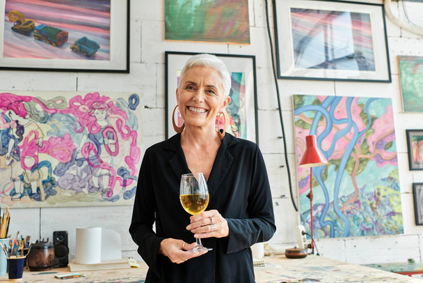 Şarap kadehiyle neşeli olgun kadın sanatçı sanat stüdyosunda yaratıcı resimlerle kameraya gülümsüyor. - Fotoğraf, Görsel