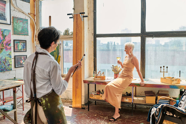 élégant femme mature posant avec verre à vin près artiste féminine dans un atelier léger et spacieux - Photo, image