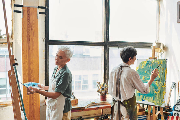 mujeres creativas de mediana edad en delantales pintura sobre caballetes en taller de artesanía moderna, hobby artístico - Foto, Imagen