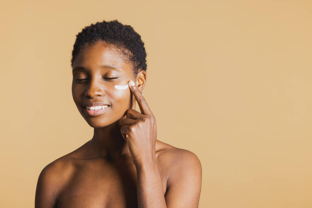 Joven mujer negra sonriente con los ojos cerrados aplicando crema hidratante en su cara. Mujer joven tratando su piel facial con un producto de belleza. - Foto, Imagen