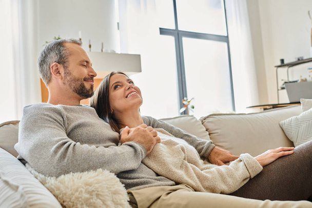 pleased child-free couple sitting on comfortable couch and looking at each other in living room - Photo, Image