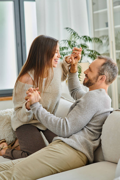 joyful child-free couple holding hands and laughing at each other on couch in living room, harmony - Photo, Image