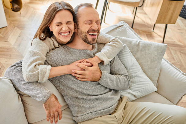 mulher animada com os olhos fechados abraçando marido rindo no sofá na sala de estar, casal sem filhos - Foto, Imagem