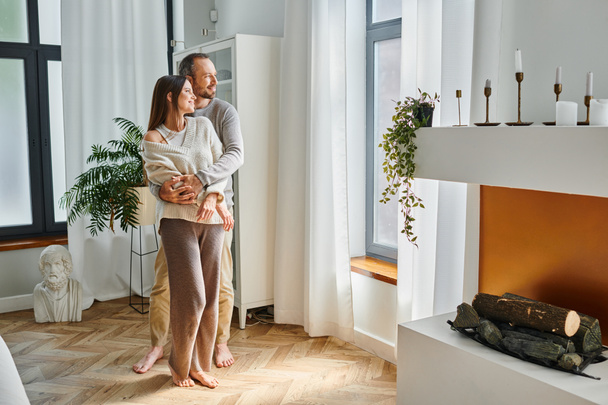 felice coppia senza bambino in piedi alla finestra in accogliente soggiorno con interni moderni e camino - Foto, immagini