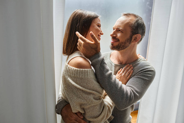 couple aimant sans enfant embrasser et sourire à l'autre près de la fenêtre à la maison, tendresse - Photo, image