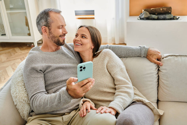 Lächelndes kinderfreies Paar mit Smartphone, das es sich zu Hause auf der Couch gemütlich macht und einander anschaut - Foto, Bild