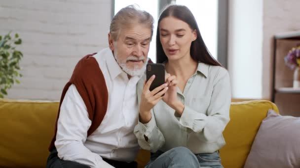 Jeune femme heureuse enseignant à son grand-père aîné à utiliser l'application mobile sur smartphone, passer du temps ensemble à la maison, regarder des vidéos et rire - Séquence, vidéo