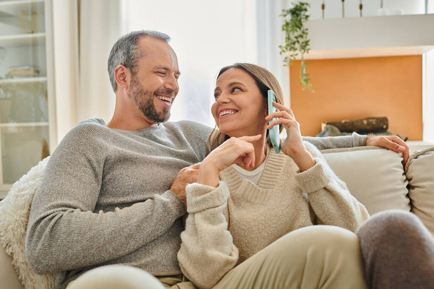 счастливая женщина разговаривает на диване смартфона в гостиной рядом с улыбающимся мужем, без детей пара - Фото, изображение