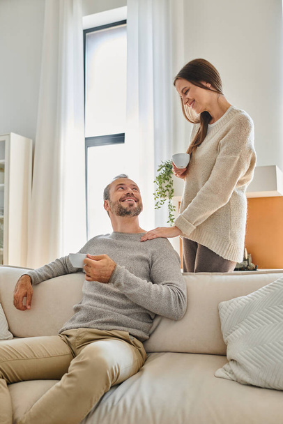 bezdětný pár s šálky kávy usmívající se na sebe v útulném obývacím pokoji doma, šťastné ráno - Fotografie, Obrázek