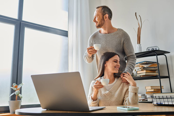 χαμογελαστός άνδρας με κούπα καφέ που υποστηρίζει τη σύζυγο που εργάζεται σε φορητό υπολογιστή στο σπίτι, ενότητα του ζευγαριού χωρίς παιδιά - Φωτογραφία, εικόνα