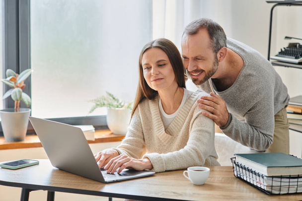 щасливий чоловік обіймає плечі усміхненої дружини, яка працює на ноутбуці вдома, бездітний спосіб життя - Фото, зображення