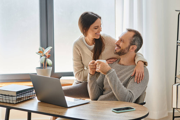 šťastný muž s šálkem kávy sedí v blízkosti notebooku a pečující manželka v domácí kanceláři, dítě-volný životní styl - Fotografie, Obrázek