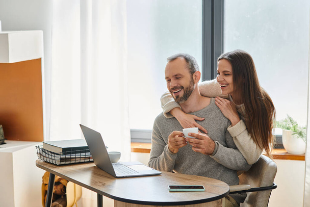 mujer alegre abrazando marido sonriente sentado con taza de café cerca de la computadora portátil, estilo de vida libre de niños - Foto, Imagen