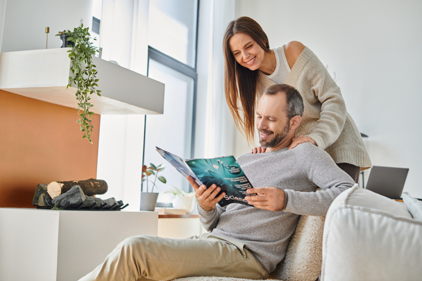 freudiger Mann liest Wissenschaftsmagazin neben lächelnder Frau auf Couch im Wohnzimmer, kinderfreies Paar - Foto, Bild