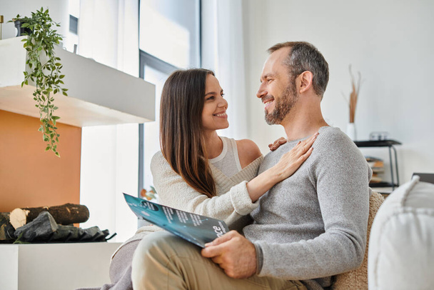smiling woman embracing husband reading science magazine on couch at home, child-free life - Photo, Image