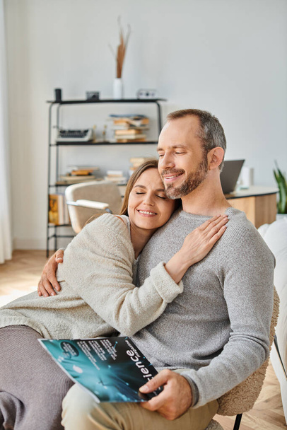 delighted woman hugging husband sitting with science magazine on couch at home, child-free life - Photo, Image