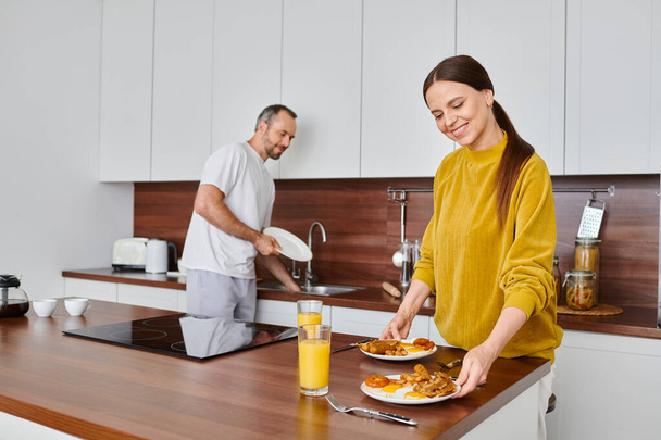 χαμογελαστή γυναίκα σερβίρει νόστιμο πρωινό, ενώ ο σύζυγος πλένει πιάτα στην κουζίνα, φροντίδα και αρμονία - Φωτογραφία, εικόνα