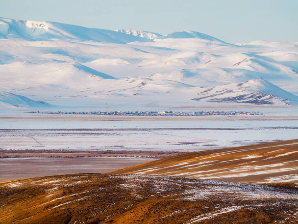 A pequena cidade autêntica na distância é coberta da neve na distância, a aldeia siberiana de Kokorya no sopé da montanha no inverno. Condições de vida difíceis no Norte. - Foto, Imagem