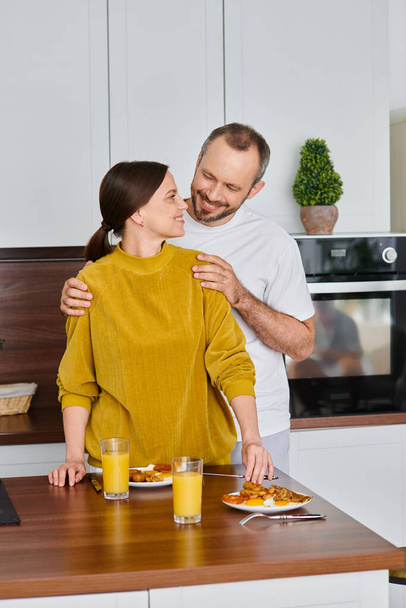 Gülümseyen adam, karısının omuzlarına sarılıp kahvaltıyı hazırlıyor, çocuksuz bir çiftin sabahında. - Fotoğraf, Görsel