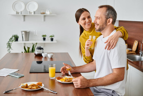 χαρούμενη σύζυγος κρατώντας χυμό πορτοκαλιού κοντά στον σύζυγο απολαμβάνοντας το πρωινό στην κουζίνα, ζευγάρι χωρίς παιδιά - Φωτογραφία, εικόνα