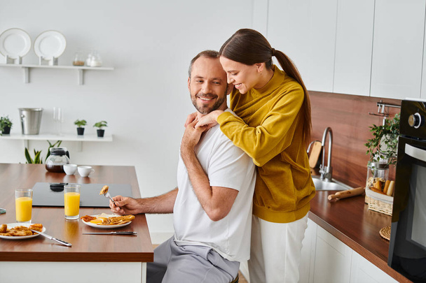widok z boku uśmiechnięty mężczyzna korzystających smaczne śniadanie w pobliżu opiekuńczej żony w kuchni, para wolna od dzieci - Zdjęcie, obraz