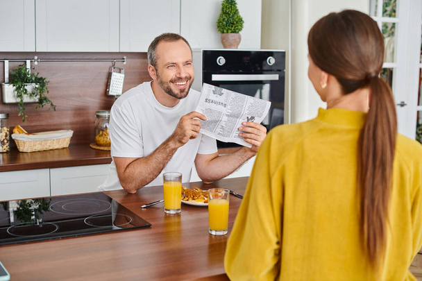χαρούμενος άνθρωπος δείχνει εφημερίδα στη σύζυγο κατά τη διάρκεια του πρωινού στη σύγχρονη κουζίνα, χωρίς παιδιά τρόπο ζωής - Φωτογραφία, εικόνα