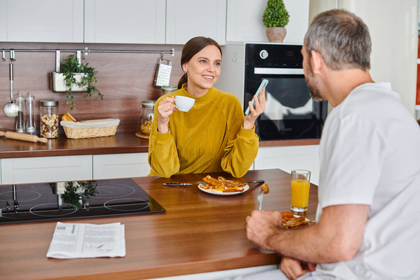 cheerful woman with coffee and smartphone talking to husband during breakfast, child-free lifestyle - Photo, Image