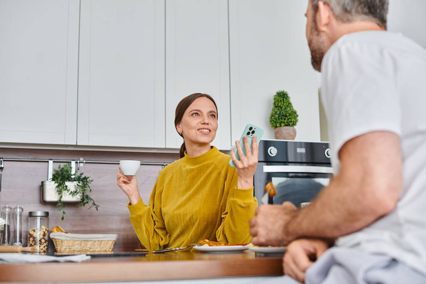взволнованная женщина со смартфоном и чашкой кофе разговаривает с мужем во время завтрака на кухне - Фото, изображение