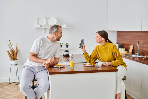 freudige Frau zeigt ihrem Mann das Smartphone beim Frühstück in der modernen Küche, kinderfreies Paar - Foto, Bild