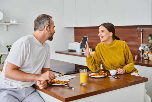 happy woman showing smartphone to husband during breakfast in modern kitchen, child-free couple - Photo, Image