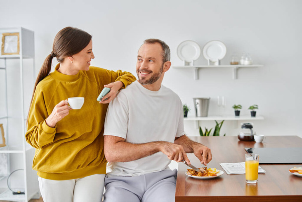 весёлый мужчина наслаждается вкусным завтраком рядом с женой с чашкой кофе и смартфоном на современной кухне - Фото, изображение