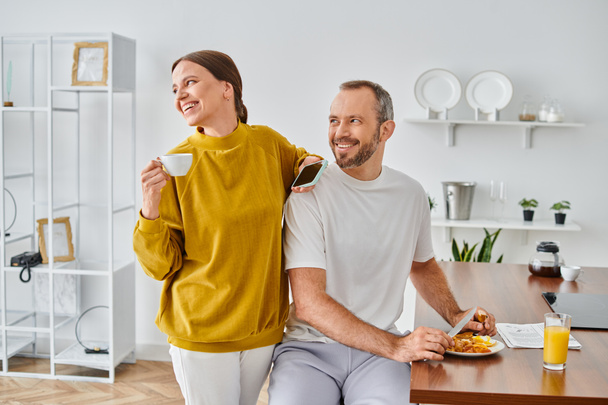 homem feliz desfrutando de saboroso café da manhã perto da esposa com xícara de café e smartphone olhando para longe na cozinha - Foto, Imagem