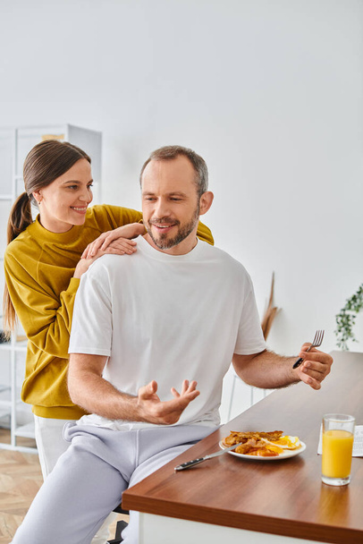 Heyecanlı adam kahvaltı yapıyor ve modern mutfakta gülümseyen eşiyle konuşuyor, çocuksuz bir çift. - Fotoğraf, Görsel