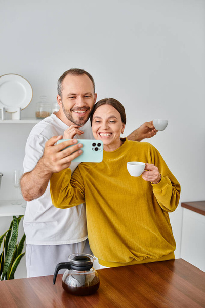 joyful child-free couple talking self-portrait on smartphone during morning coffee in kitchen - Photo, Image