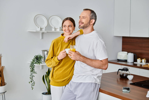 coppia senza bambini sorridente con caffè aromatico e succo d'arancia fresco guardando altrove in cucina - Foto, immagini