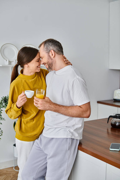 radostný bezdětný pár s aromatickou kávou a čerstvou pomerančovou šťávou, usmívající se tváří v tvář v kuchyni - Fotografie, Obrázek
