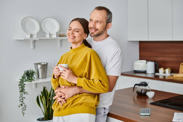 caring man embracing delighted wife holding fresh morning coffee in kitchen, child-free concept - Фото, изображение
