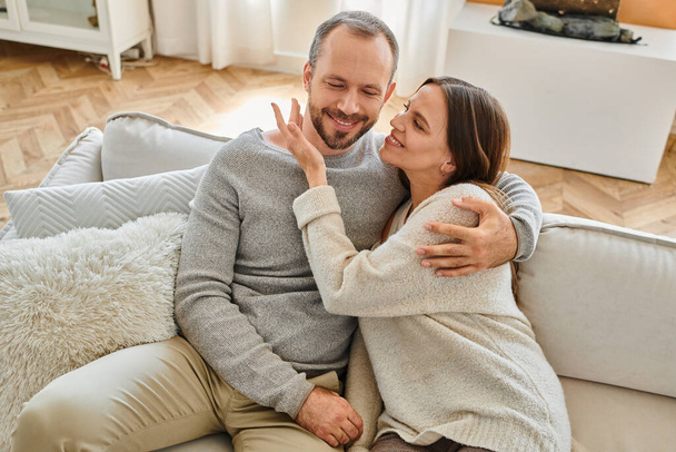 joyful woman embracing happy husband on cozy couch in living room, leisure of child-free couple - Photo, Image