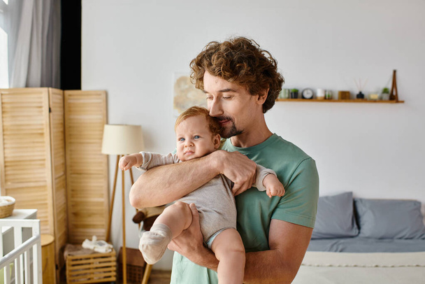 カーリー・ヘアと陽気な父親は,自宅で居心地の良い寝室で赤ちゃんの男の子を抱えています, 父親時代 - 写真・画像