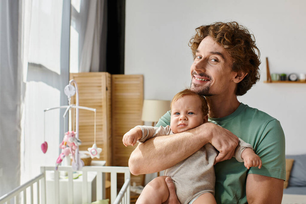 padre rizado y feliz sosteniendo en brazos a su hijo en el acogedor dormitorio en casa, la paternidad - Foto, Imagen