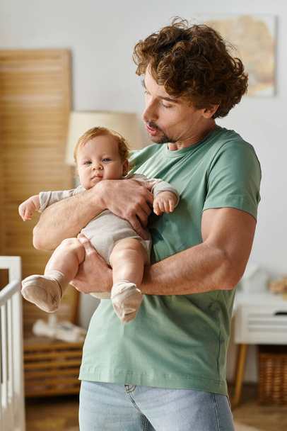彼の赤ん坊の息子を自宅で居心地の良い寝室で抱えているカーリー髪とひげ付きの男 - 写真・画像