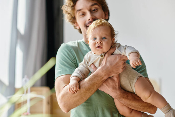 hombre de pelo rizado y barbudo sosteniendo en brazos a su hijo bebé con ojos azules en casa, paternidad - Foto, Imagen