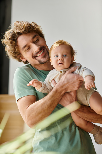 σγουρά μαλλιά και ευχαριστημένος πατέρας κρατώντας στην αγκαλιά το μικρό του γιο στο άνετο υπνοδωμάτιο στο σπίτι, πατρότητα - Φωτογραφία, εικόνα